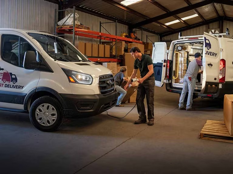 Three workers loading and organizing cardboard boxes into white delivery vans inside a warehouse.