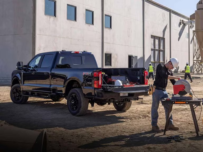 Worker in hard hat using a power saw near a black pickup truck with equipment loaded in the bed at an active construction site.